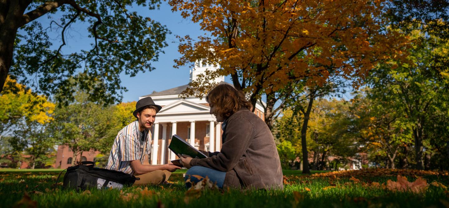 Two students study together outside under the bright colors of autumn trees.